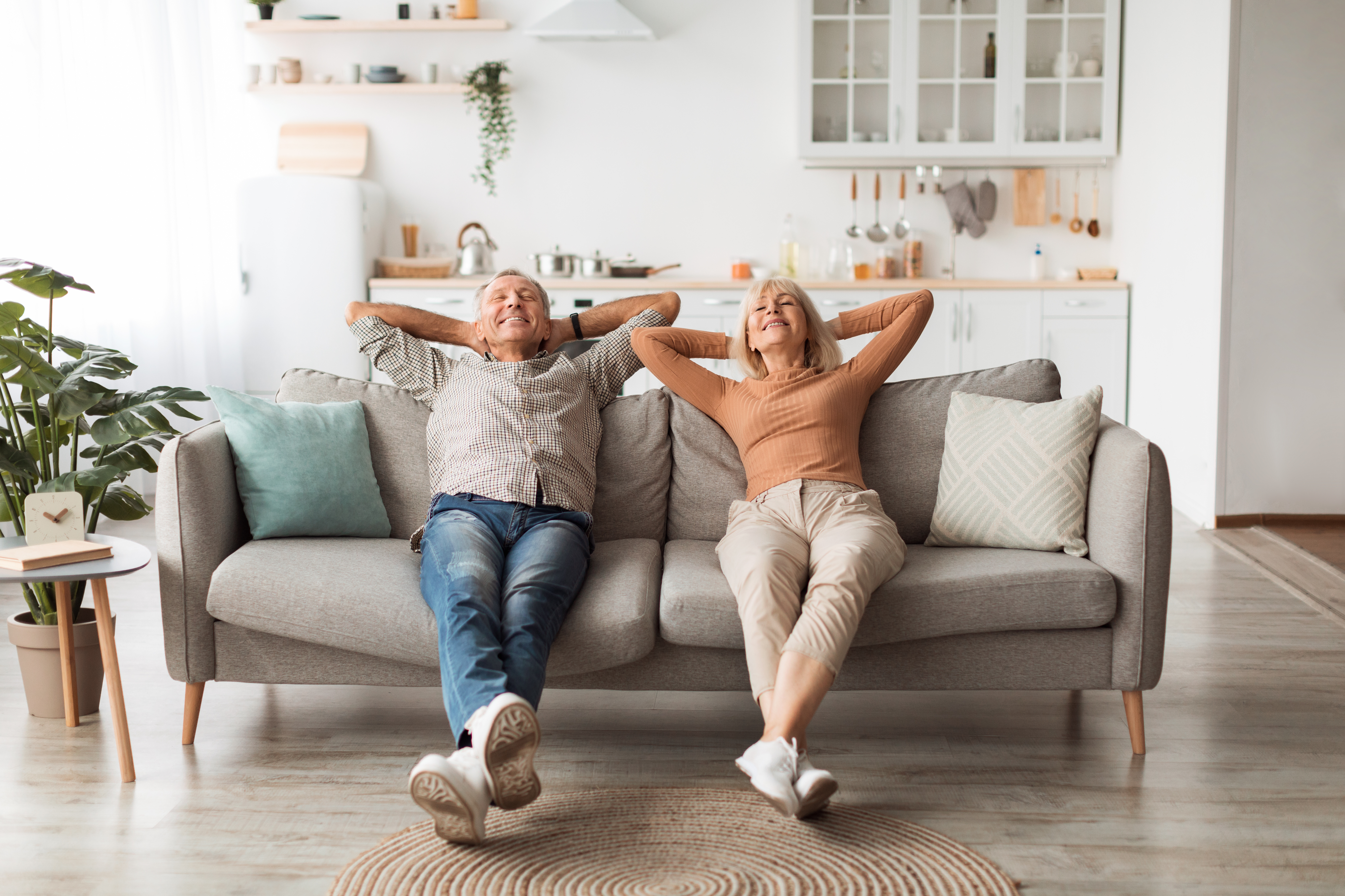 Happy Senior Couple Relaxing Sitting On Sofa Holding Hands Behind Head And Closing Eyes Enjoying Rest At Home On Weekend. Domestic Comfort And Relaxation Concept
